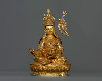 May include: A gold-colored statue of a deity sitting in a meditative pose. The statue is adorned with intricate details and a crown. The deity is holding a staff in their right hand and a hand gesture in their left hand. The statue is sitting on a lotus base.