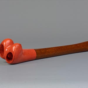 Può includere: Una pipa di legno con un fornello rosso bulboso con due aperture. La pipa è in legno e ha una finitura naturale.