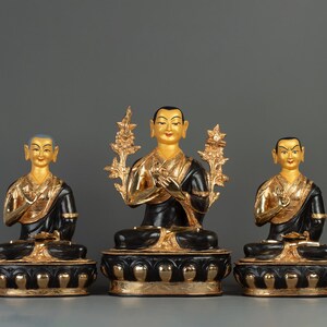 Puede incluir: Tres estatuas doradas y negras de figuras budistas sentadas en posición de meditación. La figura central sostiene una flor en su mano. Las estatuas están sobre bases negras y doradas.