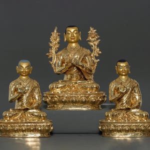 Peut inclure: Trois statues dorées de figures bouddhistes assises en position de méditation. Les statues sont ornées de détails complexes et sont placées sur des socles ornés.