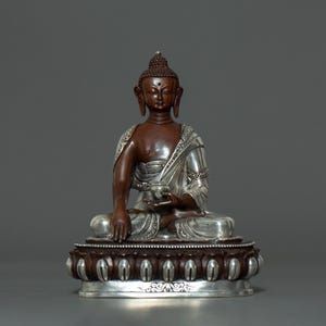 Peut inclure: Une statue de Bouddha en bronze assis avec une finition argentée. Le Bouddha tient un bol sur ses genoux et porte une robe aux détails complexes. La statue est assise sur une base de lotus.