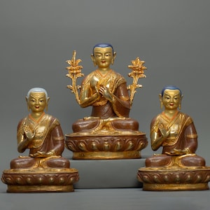 Puede incluir: Tres estatuas doradas de figuras budistas sentadas en posición de meditación. Las figuras visten ropas rojas y doradas y tienen detalles intrincados. Las estatuas están sobre un fondo gris.