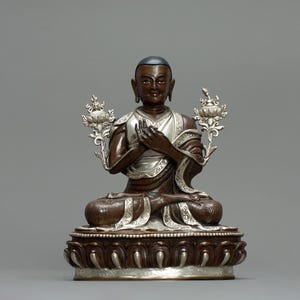 Puede incluir: Una estatua de bronce de un Buda sentado con una corona de plata y detalles ornamentados. El Buda está en una postura meditativa con las manos juntas frente al pecho.