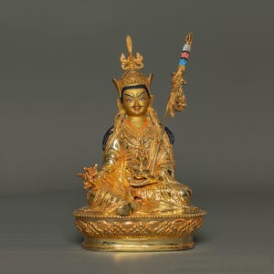 Könnte beinhalten: Eine goldene Statue einer sitzenden Figur, geschmückt mit einer detaillierten Krone und Gewändern. Die Statue hält einen Stab mit farbenfrohen Akzenten. Die Figur hat einen ruhigen Ausdruck und sitzt auf einem dekorativen Sockel. Das Gesamtdesign ist aufwendig und verziert.