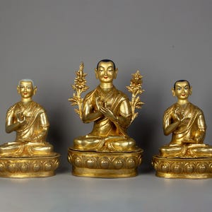 Puede incluir: Tres estatuas doradas de figuras budistas en posición de meditación sentada. Las figuras visten ropas tradicionales y tienen expresiones serenas. Las estatuas se colocan sobre bases ornamentadas con intrincados detalles.