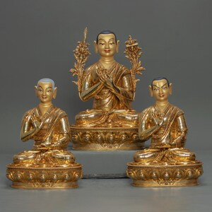 Puede incluir: Tres estatuas doradas de monjes budistas sentados en posición de meditación. Cada estatua está sentada sobre una base de loto con intrincados detalles. Las estatuas están dispuestas en fila, con la estatua central ligeramente más grande que las otras.