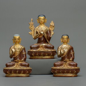 Puede incluir: Tres estatuas de bronce doradas que representan a monjes budistas sentados. Los monjes llevan túnicas marrones y tienen adornos dorados. Las estatuas están sobre un fondo gris.