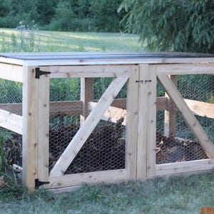 Op de afbeelding: Een houten compostbak met een lattenbodem en twee scharnierende deuren. De bak is gemaakt van lichtgekleurd hout en heeft zijkanten van gaas. Zwarte scharnieren en een grendel bevestigen de deuren. De bak staat buiten, omgeven door gras en groen.