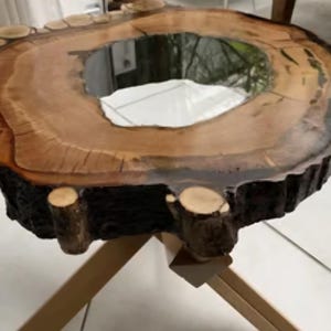 Może przedstawiać: Unikalny, ręcznie wykonany stolik kawowy z przekroju pnia drzewa. Stół ma naturalne usłojenie drewna z ciemną, zwęgloną krawędzią i przezroczystym, odbijającym światło środkiem. Stół wspiera jasna drewniana podstawa. Średnica: ok. 76 cm.