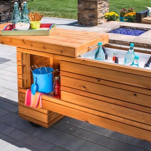 Op de afbeelding: Een houten buitenkoeler met een scharnierend tafelblad. De koeler is gevuld met ijs en drankjes. Een blauwe emmer, een schep en een rode fles zijn zichtbaar aan de zijkant. Een dienblad met snacks en drankjes staat op het tafelblad.