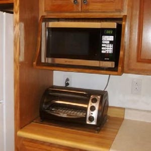 Puede incluir: Un horno microondas negro y una tostadora negra están empotrados en gabinetes de cocina de madera. El microondas tiene una pantalla digital y un panel de control. La tostadora tiene tres perillas de control y un borde plateado.