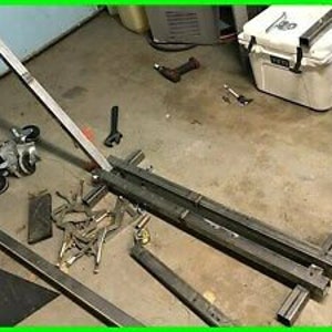 May include: A metal frame with wheels and a metal bar attached to it. The frame is made of square tubing and the bar is made of round tubing. The frame is sitting on a concrete floor.