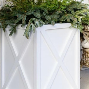 Puede incluir: Una jardinera de madera blanca con un diseño en forma de X en los lados, llena de ramas de hoja perenne frescas. La caja es cuadrada y está colocada en un porche, lo que sugiere su uso en exteriores para la decoración de temporada.