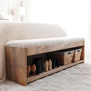 May include: A wooden bench with a light-colored cushioned top and a lower shelf. The shelf holds various shoes and two woven baskets. The bench is placed at the foot of a bed, creating a functional and stylish storage solution.