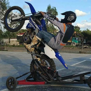 Puede incluir: Una persona realizando un wheelie en una moto de cross azul y blanca con un casco negro y equipo de conducción naranja y gris. La moto está en un soporte de metal negro con ruedas.