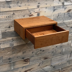 Peut inclure: Une table de chevet flottante en bois avec un tiroir ouvert. La table de chevet est en bois brun clair et est fixée à un mur en bois rustique et vieilli. Le tiroir est ouvert, révélant l'intérieur.