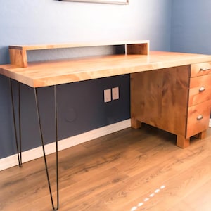 May include: A wooden desk with a light-colored top and a built-in shelf. The desk has three drawers on the right side and is supported by black hairpin legs. The desk is set against a blue wall and hardwood floor.