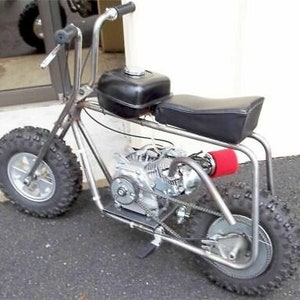 May include: A small, custom-built motorcycle with a black gas tank, a black seat, and knobby tires. The motorcycle has a single-cylinder engine and a red air filter.