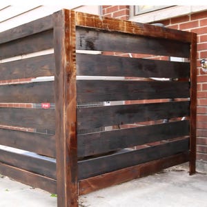 Könnte beinhalten: Ein dunkel gebeizter Sichtschutz aus Holz mit horizontalen Lamellen und einem rechteckigen Rahmen. Der Sichtschutz befindet sich vor einer Backsteinmauer. Das Holz hat eine natürliche Maserung und Textur. Ein kleines rotes Schild ist angebracht.