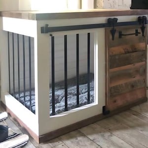 May include: A wooden dog crate with a sliding barn door. The crate has a white frame, a dark wooden top, and black metal bars. The sliding door is made of reclaimed wood. A dog bed is visible inside the crate.
