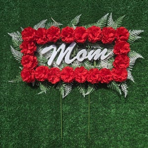 May include: A rectangular decorative sign featuring the word Mom in white script lettering, surrounded by a border of vibrant red carnations and light green fern-like foliage, supported by two thin green metal stakes extending from the bottom.