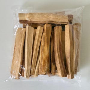 Peut inclure: Un sac en plastique transparent rempli de bâtonnets de palo santo brun clair. Les bâtonnets présentent un grain de bois naturel et différentes nuances de brun. Le sac est scellé et les bâtonnets sont regroupés.