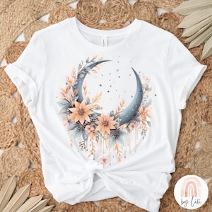Puede incluir: Camiseta blanca con una luna creciente azul rodeada de flores de melocotón y azules. La luna tiene una textura brillante y las flores tienen un efecto acuarela. El diseño está goteando pintura dorada.