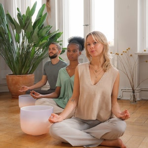 Pu&ograve; includere: Tre persone sedute in una stanza con pavimento in legno, meditando a occhi chiusi. Davanti a loro ci sono due ciotole cantanti di cristallo. Le ciotole sono di colore rosa chiaro e blu.