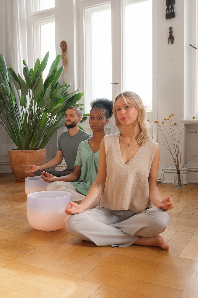 Pu&ograve; includere: Tre persone sedute in una stanza con pavimento in legno, meditando a occhi chiusi. Davanti a loro ci sono due ciotole cantanti di cristallo. Le ciotole sono di colore rosa chiaro e blu.