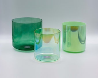 May include: Three cylindrical glass tumblers in shades of green. The glasses vary in color, with one being a deep emerald green, another iridescent, and the third a light green. The glasses are of varying heights and widths.