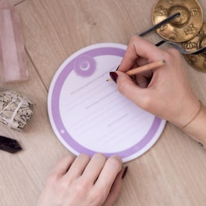 Könnte beinhalten: Ein rundes lila und weißes Journal mit linierten Seiten zum Schreiben. Eine Person schreibt mit einem Bleistift in das Journal.
