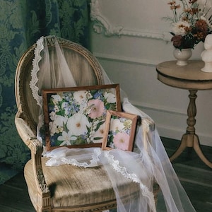 May include: A wooden chair holds two rectangular frames containing floral prints, draped with a long white lace-trimmed veil. The frames feature a natural wood finish, and the prints display a variety of white, pink, and blue flowers.