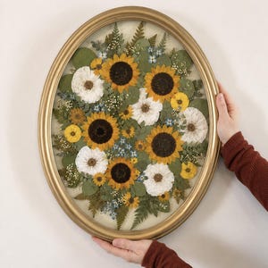 May include: An oval gold-toned frame containing a pressed floral arrangement. The composition features yellow sunflowers, white flowers, small blue blossoms, green eucalyptus leaves, and fern fronds arranged behind glass on a light background.