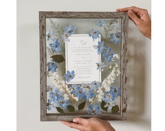 May include: A framed wedding invitation with pressed blue flowers. The invitation reads "Carly Anne & Henry Eugene Trotter III".