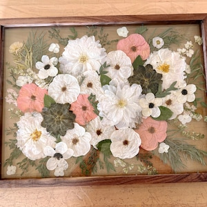May include: A rectangular wooden frame containing a collection of pressed flowers. The arrangement features white and pale pink blossoms, dark green succulent-like leaves, and thin green foliage, all preserved behind a clear glass panel.