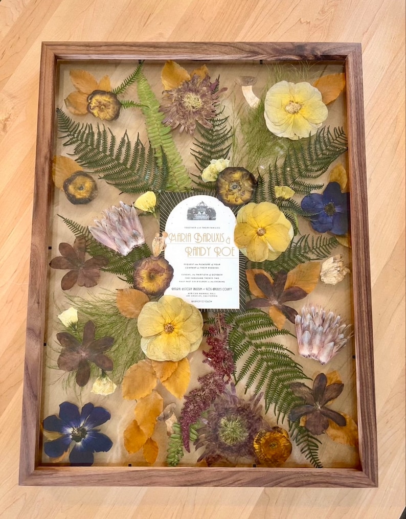 May include: A rectangular wooden frame containing a pressed flower arrangement surrounding a central wedding invitation for Maria Baruxis and Randy Roe. The display features yellow, purple, and brown dried flowers with green fern fronds.