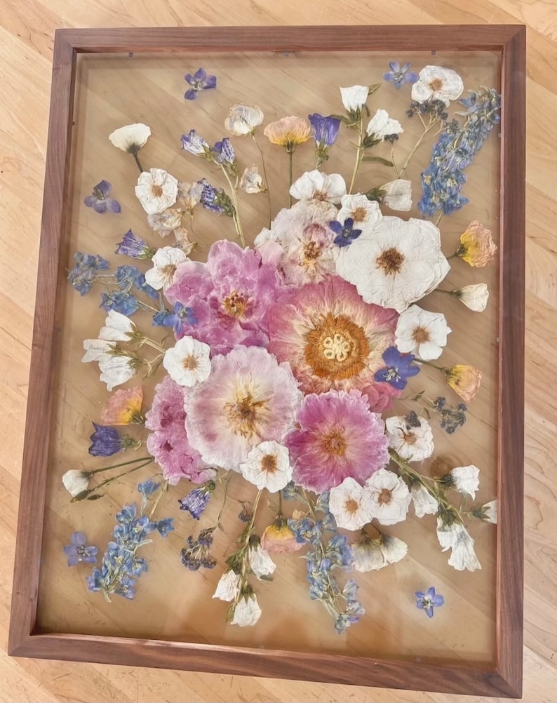May include: A rectangular wooden frame containing a pressed flower arrangement featuring pink, white, and blue blossoms. The composition includes various dried petals and stems preserved between two clear glass panes, creating a botanical display.