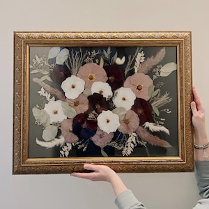 May include: A framed pressed flower arrangement with white, pink, and burgundy flowers. The flowers are arranged in a bouquet shape and are surrounded by dried grasses and leaves. The frame is gold and has a decorative border.