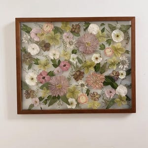 May include: A rectangular wooden frame containing a collection of pressed dried flowers in shades of pink, white, pale yellow, and green. The arrangement features various blossoms and foliage preserved behind glass, creating a botanical display.