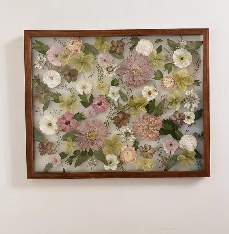 May include: A rectangular wooden frame containing a collection of pressed dried flowers in shades of pink, white, pale yellow, and green. The arrangement features various blossoms and foliage preserved behind glass, creating a botanical display.