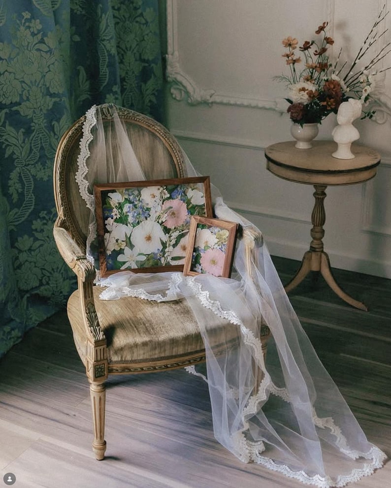 May include: A wooden chair holds two rectangular frames containing floral prints, draped with a long white lace-trimmed veil. The frames feature a natural wood finish, and the prints display a variety of white, pink, and blue flowers.