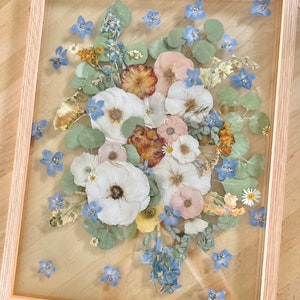 May include: A rectangular light-toned wooden frame containing a pressed flower arrangement. The display features white, pink, and yellow dried flowers interspersed with blue blossoms and green eucalyptus leaves, all set behind a clear glass pane.