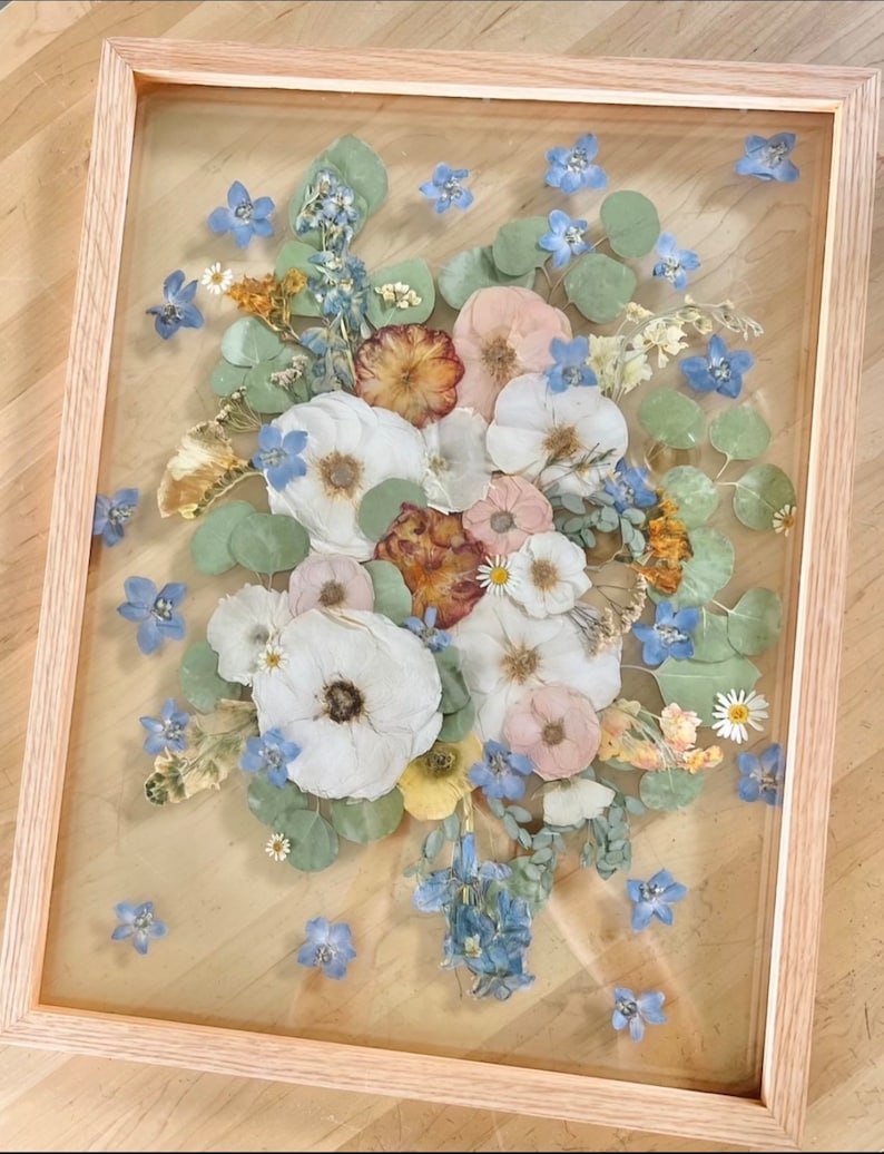 May include: A rectangular light-toned wooden frame containing a pressed flower arrangement. The display features white, pink, and yellow dried flowers interspersed with blue blossoms and green eucalyptus leaves, all set behind a clear glass pane.