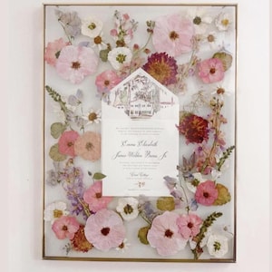May include: A framed pressed flower arrangement with a wedding invitation card inside. The invitation card is for Emma Elizabeth and James Weldon Barnes Jr. The card is decorated with a floral pattern and a small illustration of a house.