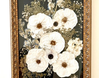 May include: A framed pressed flower arrangement with white flowers and greenery on a black background. The frame is gold with an ornate design.
