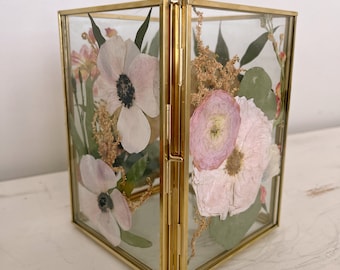 May include: A gold-toned glass box with a hinged lid, filled with pressed pink and white flowers and greenery.