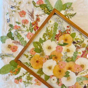 May include: A framed pressed flower arrangement with a variety of white, peach, and orange flowers, along with green leaves and stems.