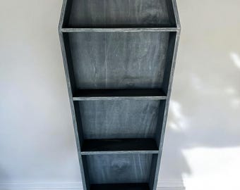 May include: A tall, coffin-shaped wooden shelving unit with a weathered gray finish. The piece features five horizontal shelves of varying widths, creating a tapered silhouette that narrows toward the bottom, constructed from wood with visible grain.