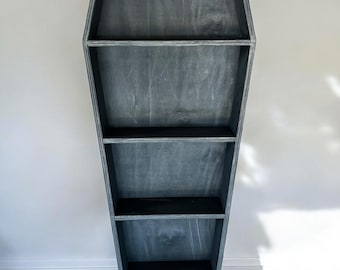 Può includere: Un alto scaffale in legno a forma di bara con una finitura grigia usurata. Il mobile presenta cinque ripiani orizzontali di larghezza variabile, creando una silhouette affusolata che si restringe verso il basso, realizzato in legno con venature.