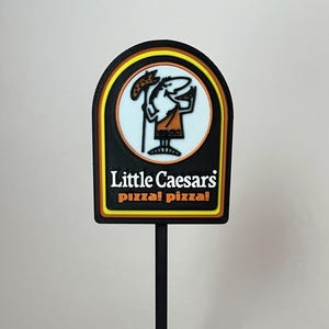 May include: A black and yellow Little Caesars pizza sign with a black stand and base. The sign features the Little Caesars logo with the text "Little Caesars pizza! pizza!". The logo includes a cartoon character holding a pizza.